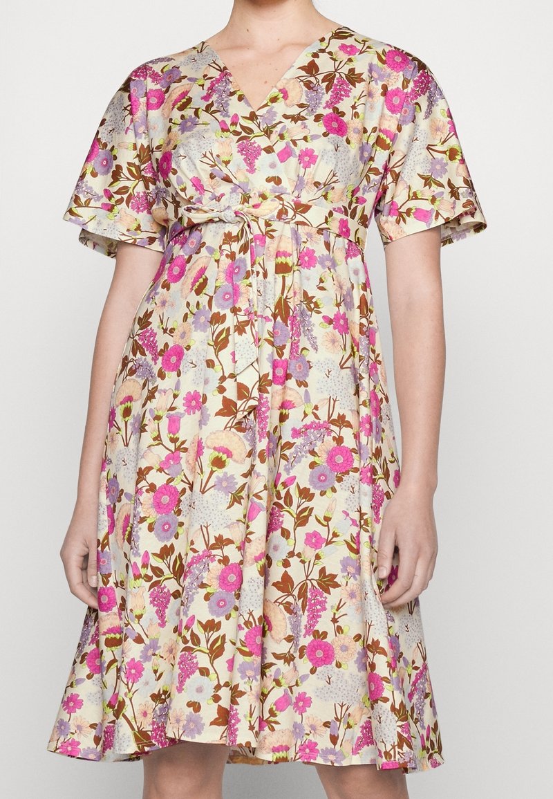 Femme portant une robe à manches courtes, longueur genou, avec un motif floral rose, violet et vert sur un fond beige clair, et une ceinture nouée à la taille.