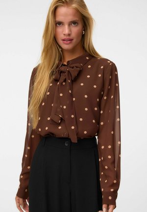 Kvinde iført en brun gennemsigtig bluse med beige polkaprikker og en stor sløjfehals, kombineret med sorte højtaljede bukser.