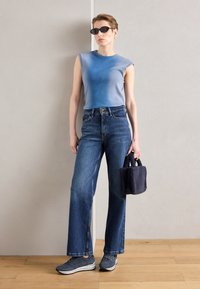 Top en mailles bleu ajusté, jeans en denim foncé taille haute avec des jambes larges, baskets grises, et un petit sac à main noir en fourrure.