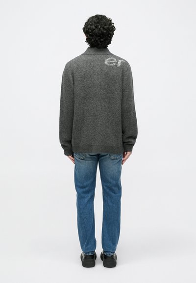 Pilkas megztas megztinis su aukštu apykakle, nugaroje su šviesiai pilkomis raidėmis „er“. Derinamas su mėlynais džinsais.