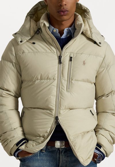 Beige puffer jacket with a hood, featuring zippered pockets and a logo on the chest. Textured fabric with a quilted pattern and adjustable closures.
