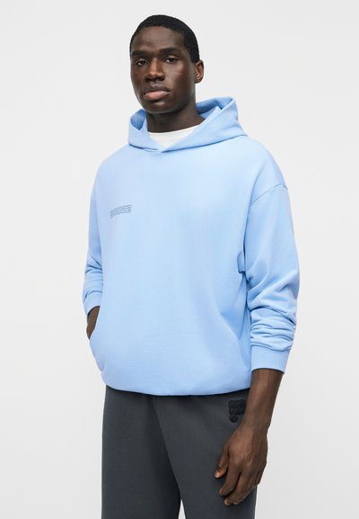 Muž v světle modré mikině s kapucí a malým textem na hrudi a tmavě šedých kalhotách, stojící s rukama v kapsách.