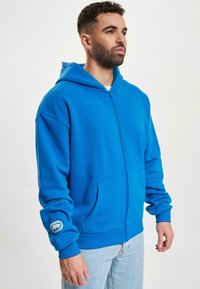 Hoodie bleu zippé en tissu doux, avec une poche avant et un logo sur le poignet gauche. Le modèle est associé à un jean en denim clair.