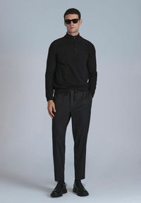 Schwarzer Strickpullover mit Reißverschlusskragen, kombiniert mit dunklen Hosen und schwarzen Schnürschuhen. Das Model trägt Sonnenbrillen und steht vor einem neutralen Hintergrund.