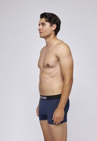 Blå boxershorts i mjukt tyg med svart elastiskt midjeband som har vit märketext. Sido vy som framhäver silhuetten.