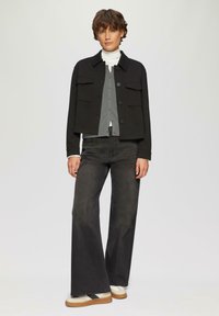 Korte zwarte jas met twee borstzakken, gelaagd over een grijze cardigan en een witte coltrui, gecombineerd met een licht uitlopende donkere spijkerbroek en sneakers.