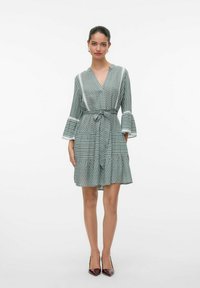 Vestido verde con estampado, escote en V, mangas acampanadas, cintura ceñida y falda acampanada. Presenta diseños geométricos en tonos claros y oscuros y un cinturón de lazo.
