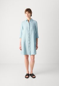 Ανοιχτό μπλε, φόρεμα πουκαμίσα μέχρι το γόνατο με κουμπιά μπροστά, τσέπη στο στήθος και μανίκια που γυρίζουν. Συνδυασμένο με μαύες παντόφλες-slide.
