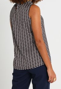 Ärmelloses Top in Schwarz mit einem geometrischen weißen Muster. Leichter Stoff mit einem dezenten Glanz. Mit einem Schlüssel-ausschnitt hinten zur Schließung.