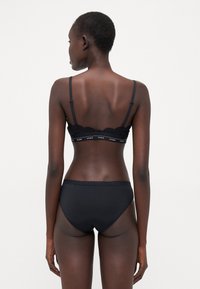 Reggiseno in pizzo nero con slip abbinati a vita alta. Il reggiseno presenta spalline regolabili e una fascia con logo, con dettagli in pizzo scallop lungo i bordi.