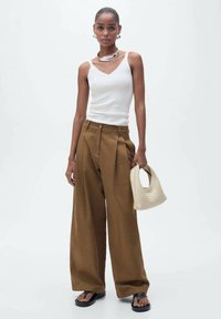 Pantaloni larghi marroni con pieghe frontali, abbinati a un top bianco senza maniche. Gli accessori includono una borsa intrecciata beige e sandali neri.