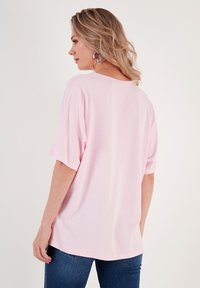 Světle růžové oversized tričko s krátkým rukávem, kulatým výstřihem a hladkým povrchem, v kombinaci s tmavě modrými džíny.
