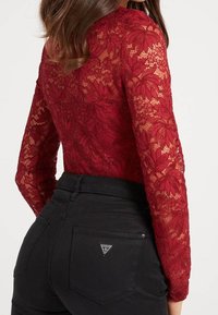 Top en dentelle rouge avec un motif floral, associé à un jean noir ajusté comportant un patch en forme de triangle sur la poche arrière.