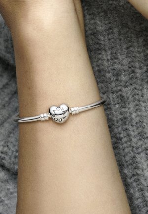 Silberarmband mit einem herzförmigen Charm, auf dem das Wort "Pandora" eingraviert ist. Glatte Textur, runde Form, mit metallischen Akzenten und hochglänzender Oberfläche.