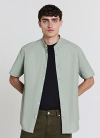Chemise à manches courtes en tissu vert clair, avec col montant et boutonnage frontal, portée sur un t-shirt noir. Design décontracté, coupe ample.