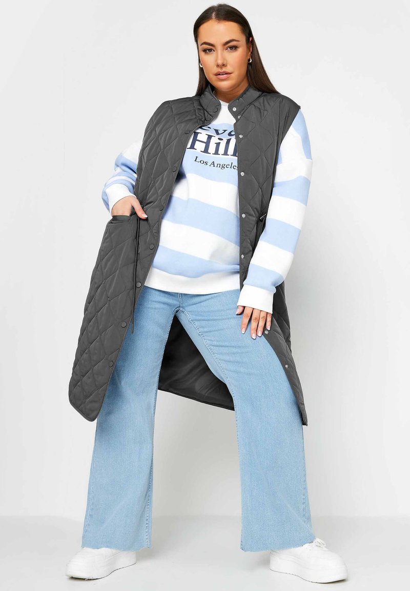 Gilet grigio trapuntato con bottoni a scatto, indossato sopra una felpa a righe blu e bianche, abbinato a jeans chiari a zampa d'elefante e sneakers bianche.