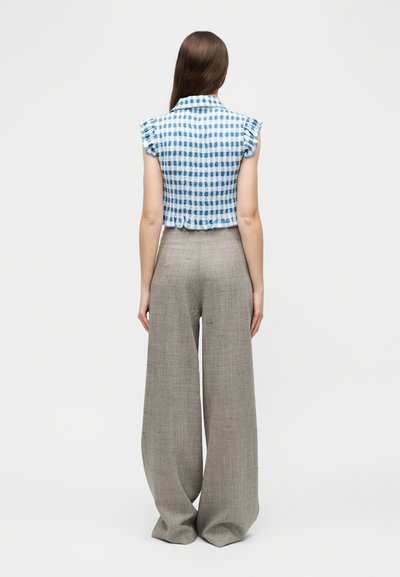 Γυναίκα με μακριά καστανά μαλλιά στέκεται με την πλάτη, φοράει μπλε και λευκό καρω μπλουζάκι χωρίς μανίκια και μπεζ παντελόνι με φαρδιά πόδια.