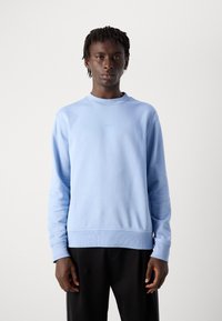 Joven con el cabello corto y enroscado, vistiendo un sudadera azul clara con el texto "BOSS" y pantalones negros, de pie contra un fondo blanco liso.