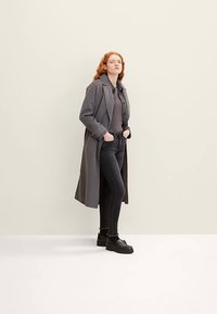 Pikk hall mantel tumeda särgi kohal, kombineeritud mustade kitsaste teksade ja mustade platvormkingadega. Siledad tekstuurid ja minimaalsed disainielemendid.