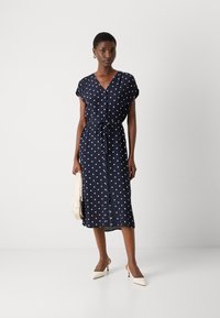 Navy polka dot dress with short sleeves, V-neckline, and tie waist detail. Fabric appears soft; knee-length design. Model holds a cream handbag.