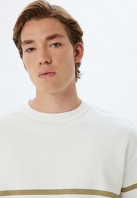 Sudadera blanca con cuello redondo, tela texturizada y una franja horizontal dorada en el pecho. Sin patrones o acentos distintivos.