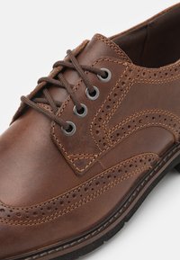 Zapato de vestir de cuero marrón con detalles de brogue, puntera redonda, seis ojales plateados y cordones marrones. Presenta costuras en contraste y una superficie texturizada.