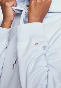 Sudadera azul claro con cordón, que presenta un pequeño logo en rojo, blanco y azul en la manga izquierda. Tejido suave y corte relajado.