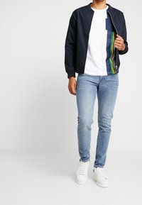 Námořnická bomber bunda, bílé tričko s modrými a zelenými pruhy, světlé modré džíny a bílé tenisky. Moderní, ležérní outfit s různými texturami.