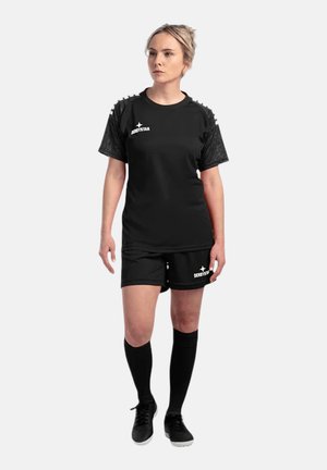Frau steht in schwarzer Sportbekleidung mit Shorts, hohen schwarzen Socken und schwarzen Schuhen vor einfarbigem Hintergrund.