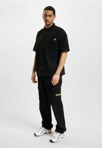 Black short-sleeve shirt with a chest pocket and green logo, paired with black cargo pants featuring side pockets and yellow embroidery. White sneakers.