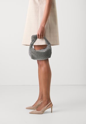 Sac à main noir et argent orné de perles avec une anse nouée, texture douce, forme arrondie. Le modèle porte une jupe beige à carreaux et des talons hauts couleur nude.