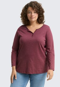 Vrouw met krullend haar, gekleed in een bordeauxrood geblokt overhemd met lange mouwen en lichtblauwe jeans, staand tegen een effen achtergrond.