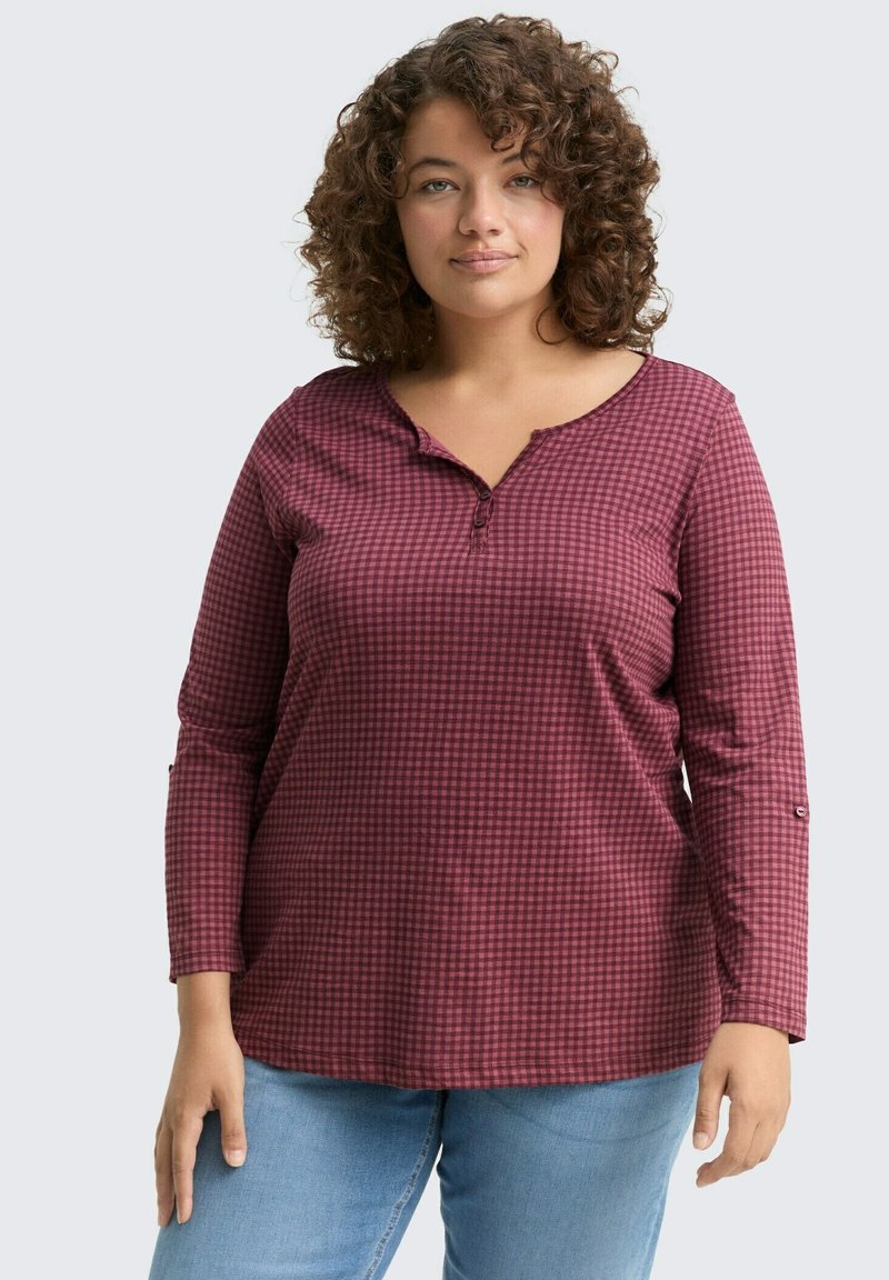 Vrouw met krullend haar, gekleed in een bordeauxrood geblokt overhemd met lange mouwen en lichtblauwe jeans, staand tegen een effen achtergrond.