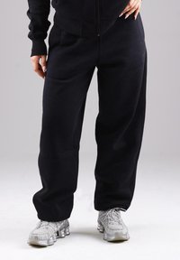 Person wearing loose black sweatpants and silver athletic shoes, standing against a plain light grey background.