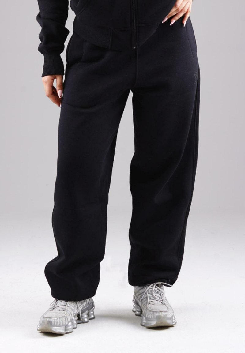 Person wearing loose black sweatpants and silver athletic shoes, standing against a plain light grey background.