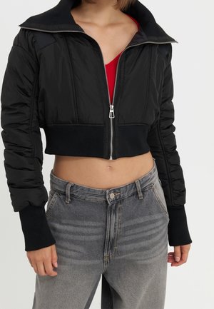 Kobieta nosząca krótka czarną kurtkę na zamek błyskawiczny, pod którą ma czerwony top oraz wysokie, wyblakłe szare dżinsy, odsłaniająca brzuch i trzymająca ręce swobodnie po bokach.