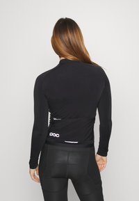 Maillot de cyclisme noir à manches longues en tissu évacuant l'humidité, coupe slim, avec un petit logo blanc à l'arrière ; associé à un short de cyclisme noir.