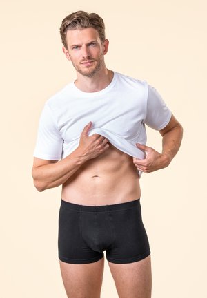 Mann in schwarzen Boxershorts hebt weißes T-Shirt, um einen durchtrainierten Bauch vor beigem Hintergrund zu zeigen.