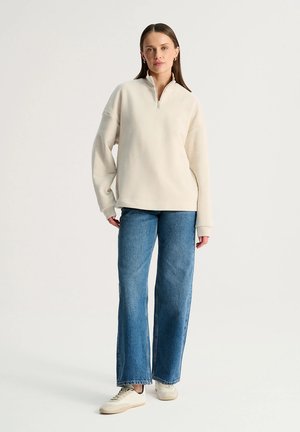 Kobieta stojąca, ubrana w kremową bluzę z ćwiartkowym zamkiem, niebieskie szerokie jeansy, białe trampki i złote kolczyki-koła na jednolitym tle.