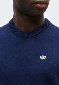 Άνδρας που φοράει μπλε πουλόβερ με στρογγυλή λαιμόκοψη, ριμπ ύφανση στη λαιμόκοψη και μικρό λευκό λογότυπο Adidas trefoil στο στήθος.
