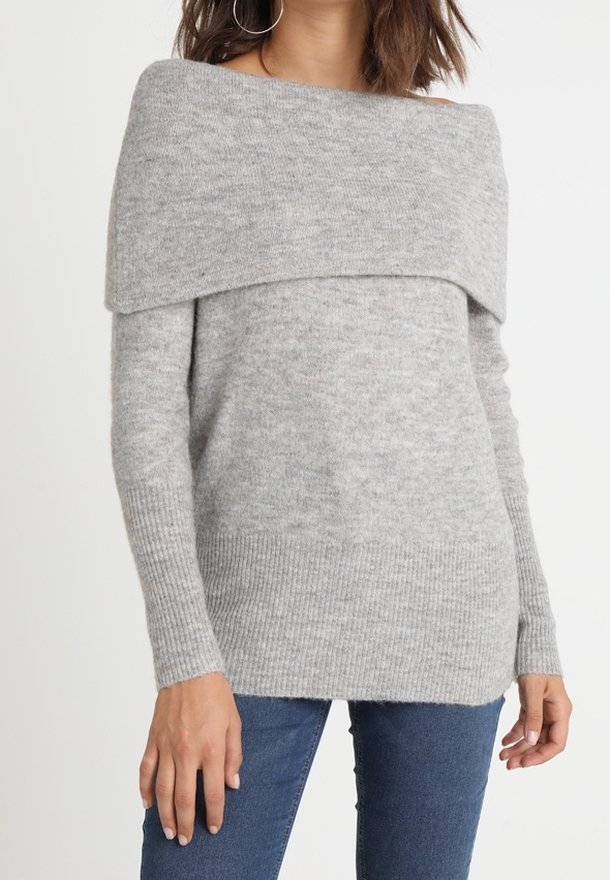 Femme portant un pull en maille gris clair à épaules dénudées avec manches longues et un jean bleu sur un fond uni.