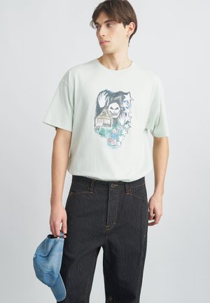 Svetlozelené bavlnené tričko s grafickým dizajnom zobrazujúcim lebku, dom a hviezdy. Kombinované s čiernymi pruhovanými nohavicami a drží modrú šiltovku.