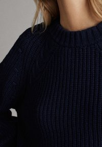 Paksu tummansininen neulepaita, jossa on ribbikuviointi, pyöreä pääntie ja kaapelitekniikkakuviointi olkapäillä.