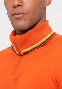 Oranžová polokošeľa s vysokým golierom, zdobená námorníckymi, žltými a červenými pruhmi. Mäkká textúra a zips prask na krku.