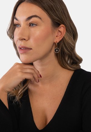 Woman with light brown hair wears black top and round gold earrings with black and gold star design, resting chin on hand.