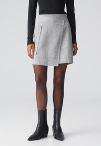 Grauer Tweed-Minirock mit Wickeldesign, ausgestattet mit seitlichen Taschen und glatter Textur, kombiniert mit schwarzen Leder-Ankle-Boots.