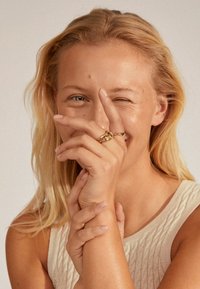 Gouden ringen met verweven ontwerpen, gedragen op de vingers. Het model heeft golvend, blond haar en poseert met een speelse uitdrukking.