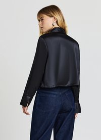 Schwarze Satinjacke mit einer kurzen Silhouette, ausgestattet mit strukturierten schwarzen Bündchen, langen Ärmeln und einem glatten Finish. Kombiniert mit dunklen Denimhose.