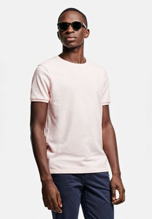 T-shirt en coton rose clair avec un col rond et des manches courtes, associée à des lunettes de soleil rondes noires et un pantalon bleu marine.