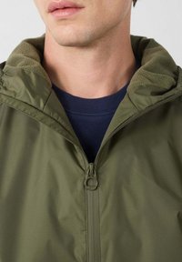 Giacca verde oliva con chiusura frontale con zip, colletto morbido foderato in pile e una camicia blu navy sotto. Tessuto liscio e strutturato.
